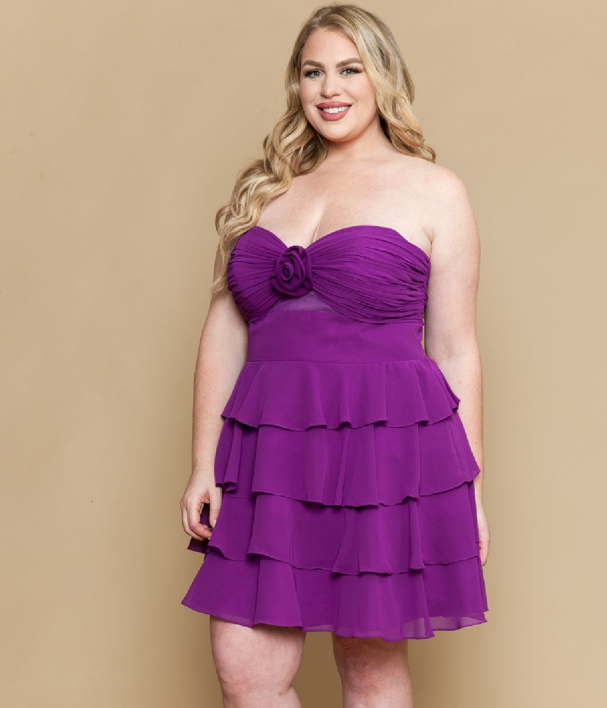 A woman smiles facing forward in the Plus-Size Short Ruffle Party Dress: Poly USA W1218 in purple, featuring tiered ruffles and a rose detail at the bust, set against a plain beige background.